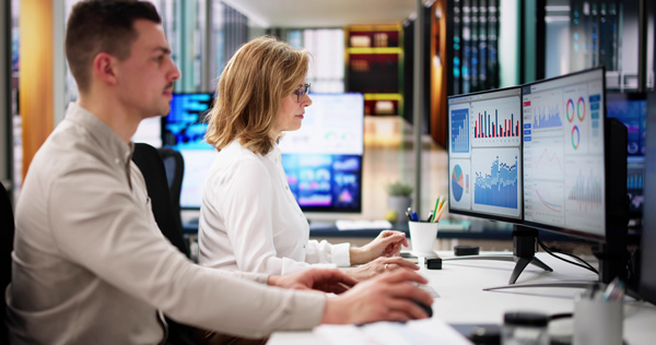 Especialistas en data analyst mujer rubia y hombre joven comparten espacio de trabajo frente a informes de analítica web en sendas pantallas de ordenador